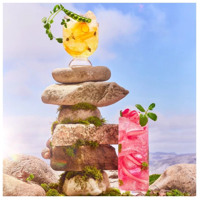 Two vibrant cocktails are balanced on a mossy rock cairn under a blue sky, evoking fresh, natural, and refreshing beverage photography.