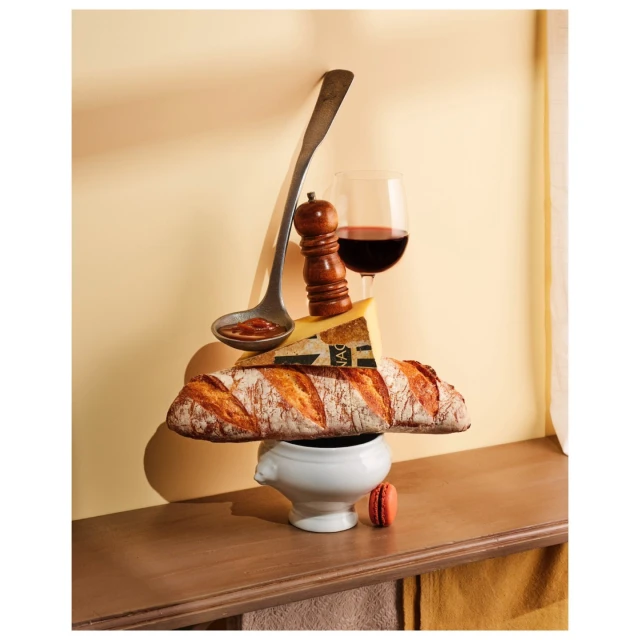 Still life with baguette, cheese, wine, and ladle creating a culinary tower on a wooden surface in a warm-toned indoor setting.