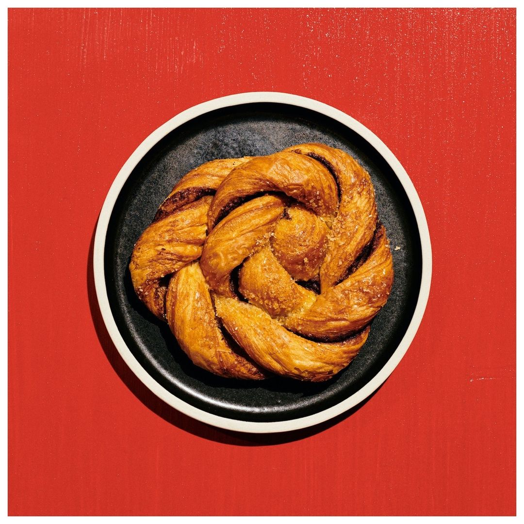 Overhead shot of a golden, twisted pastry resting on a black plate with a white rim, set against a solid, bright red backdrop.