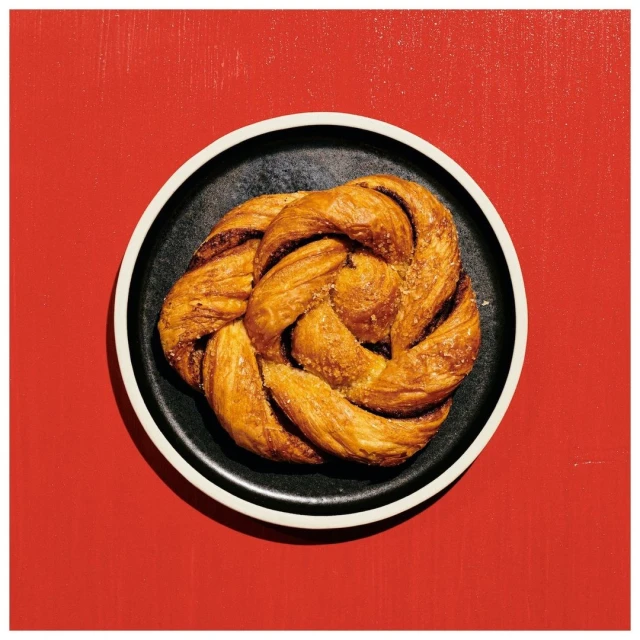 Overhead shot of a golden, twisted pastry resting on a black plate with a white rim, set against a solid, bright red backdrop.