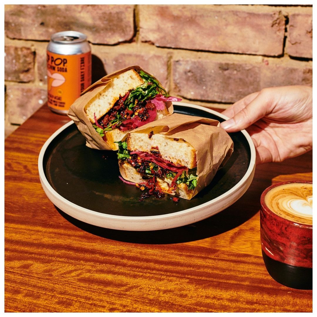 Overhead shot of a sandwich cut in half on a black plate with a soda and coffee, resting on a wooden table near a brick wall.