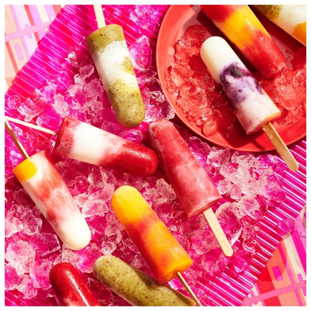 Assorted colorful popsicles on a pink plate of crushed ice and a pink background create a summery food and beverage still life.