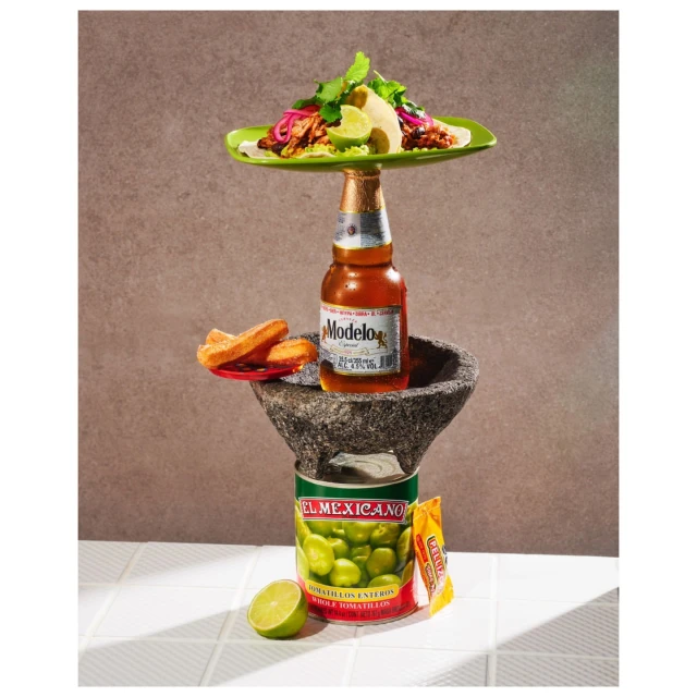 Still life with a beer bottle topped by a plate of tacos, supported by a stone mortar, a can of Mexican ingredients, and a lime.