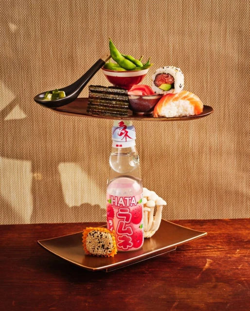 Sushi, edamame, and Japanese soda arranged on tiered trays, displayed on a wooden table against a textured beige background.