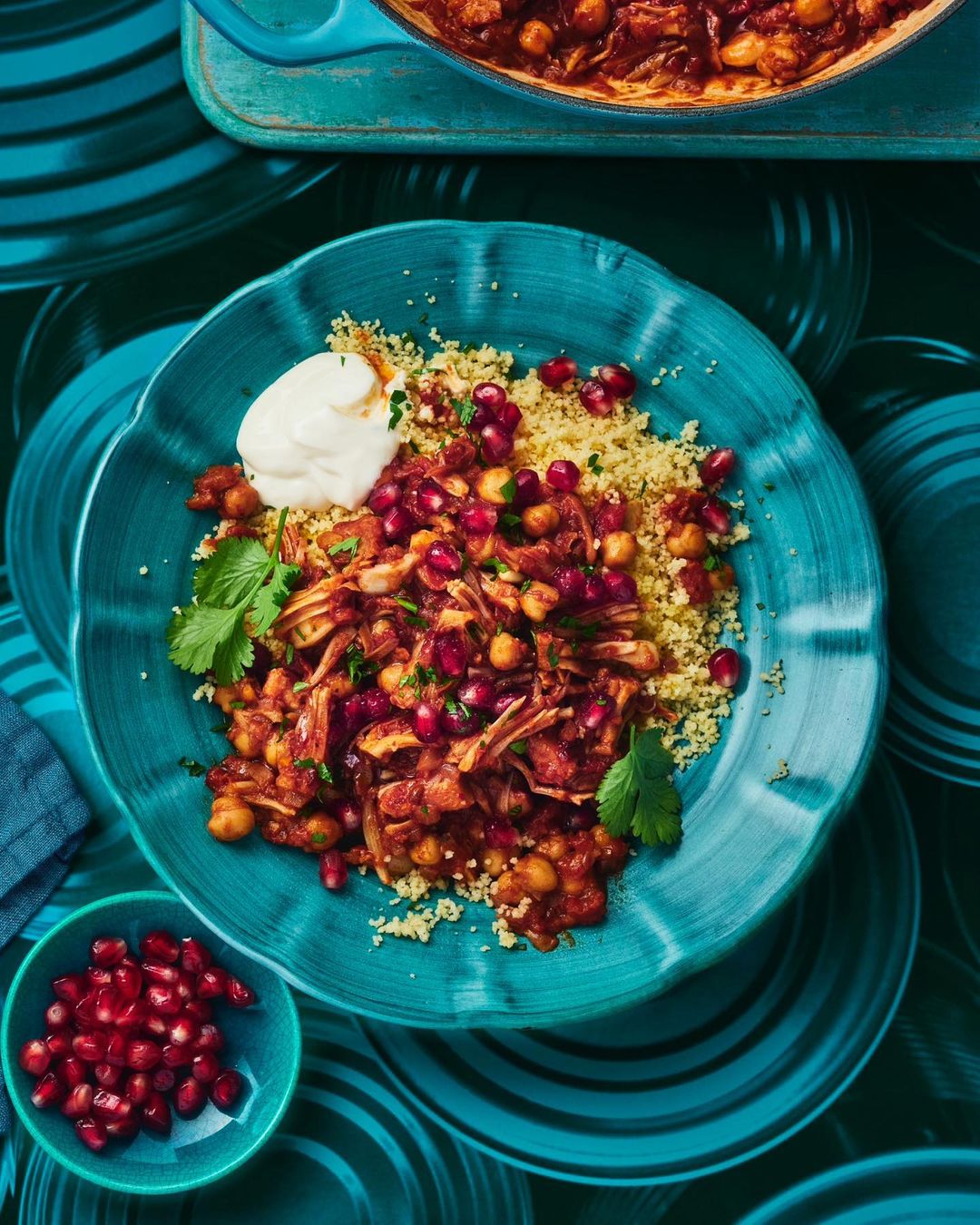 Overhead shot of chickpea stew with couscous, pomegranate seeds, and yogurt on a blue plate, creating a vibrant food composition.
