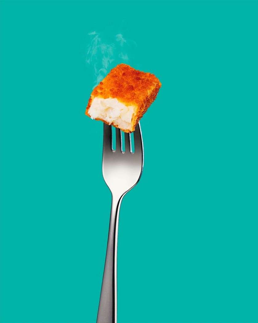 Close-up of a crispy breaded fish stick on a silver fork against a solid turquoise background with faint wisps of vapor rising.