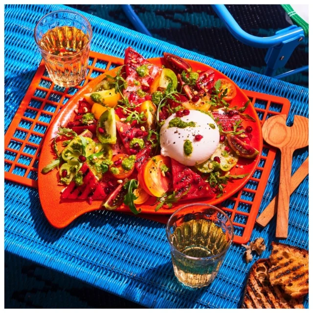 Overhead shot of caprese salad with sliced tomatoes, burrata cheese, pesto, and toast on a blue woven surface with wine glasses.