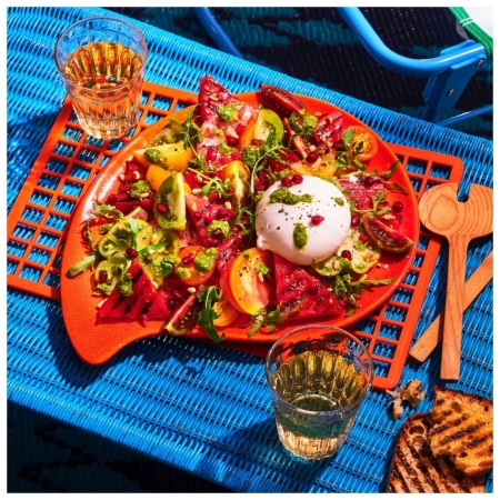 Overhead shot of caprese salad with sliced tomatoes, burrata cheese, pesto, and toast on a blue woven surface with wine glasses.