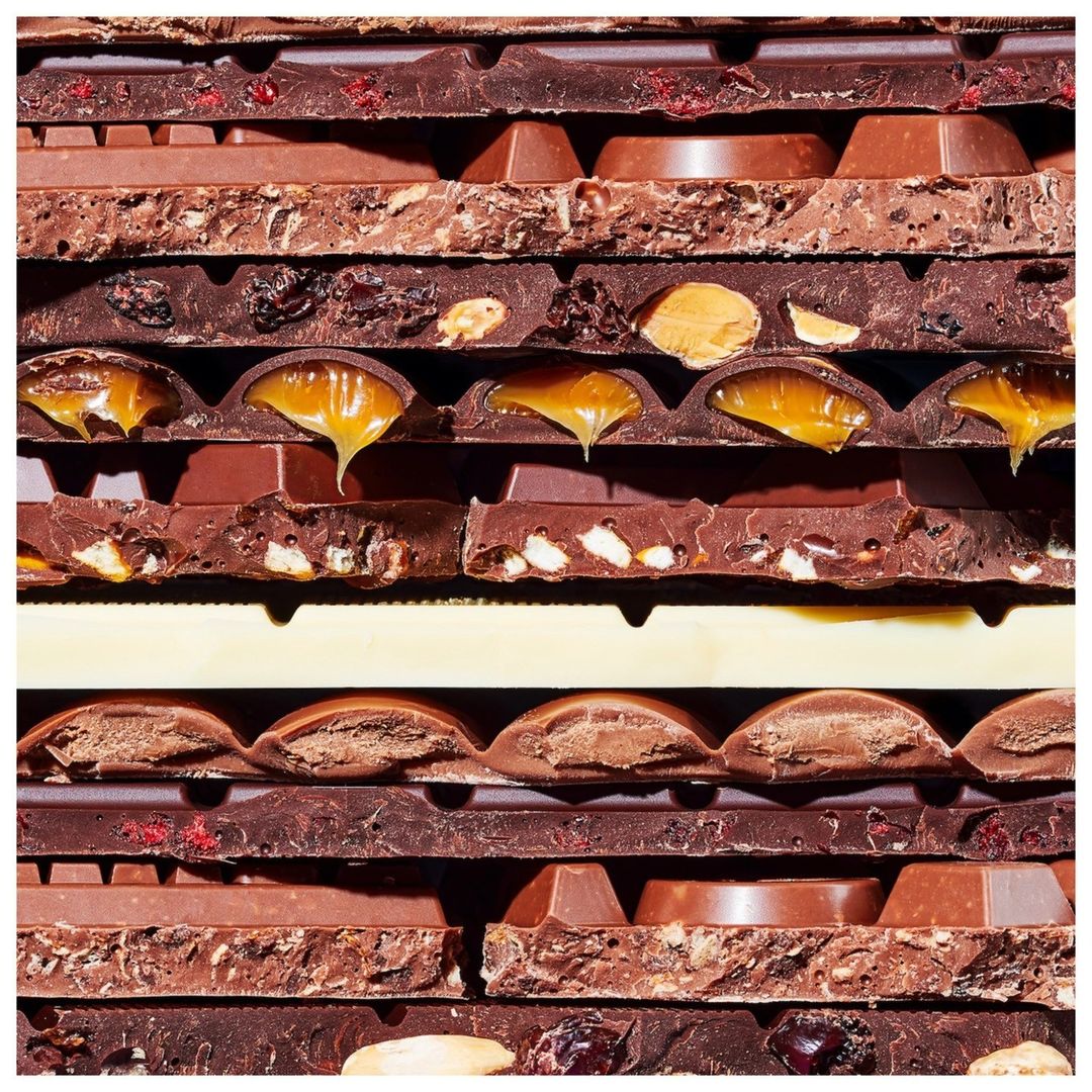 Close-up of a stack of assorted milk chocolate bars with various fillings like nuts, caramel, and dried fruits, creating a textured pattern.