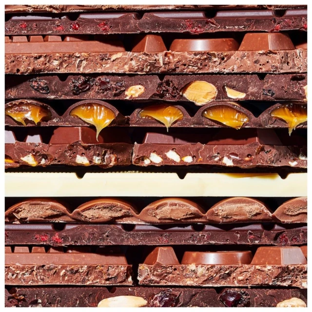 Close-up of a stack of assorted milk chocolate bars with various fillings like nuts, caramel, and dried fruits, creating a textured pattern.