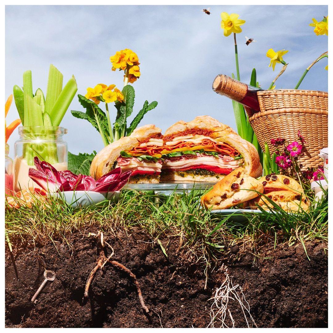 Overhead shot of a sliced sandwich loaf on a glass plate, surrounded by vegetables, fruit, a bottle of wine, and flowers in a grassy setting.