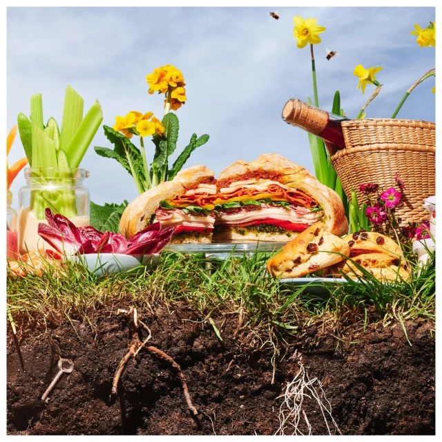 Overhead shot of a sliced sandwich loaf on a glass plate, surrounded by vegetables, fruit, a bottle of wine, and flowers in a grassy setting.