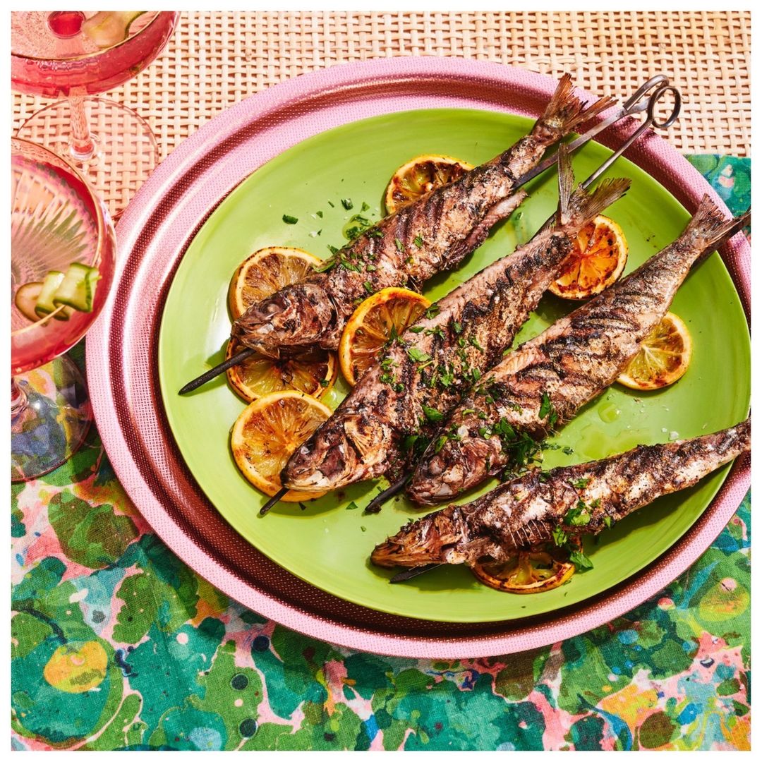 Overhead shot of grilled fish skewers with lemon slices on a green plate. Pink tray, floral tablecloth, and glass add colorful detail.