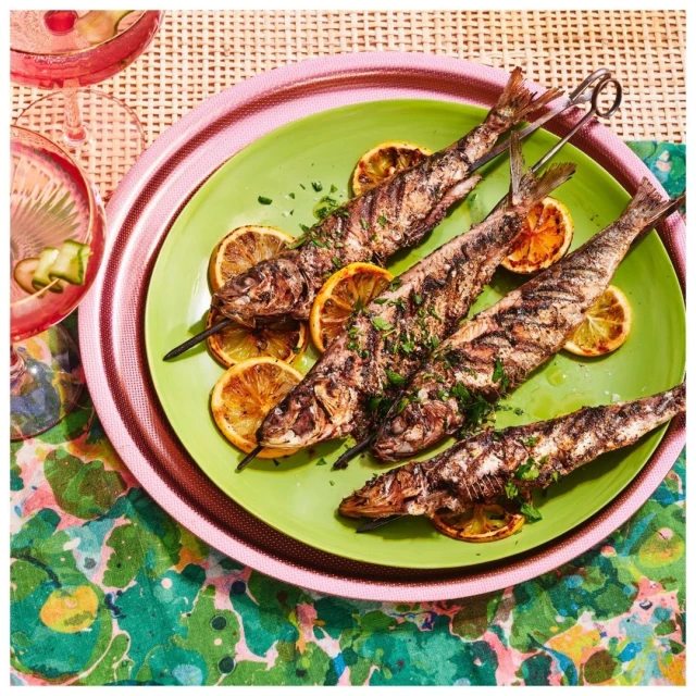 Overhead shot of grilled fish skewers with lemon slices on a green plate. Pink tray, floral tablecloth, and glass add colorful detail.