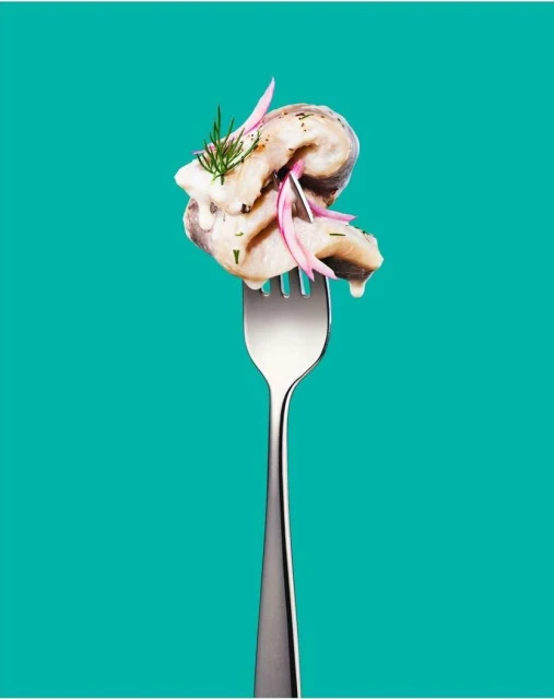 Close-up of marinated herring on a fork with dill and sliced red onion against a vibrant turquoise background, studio lighting.