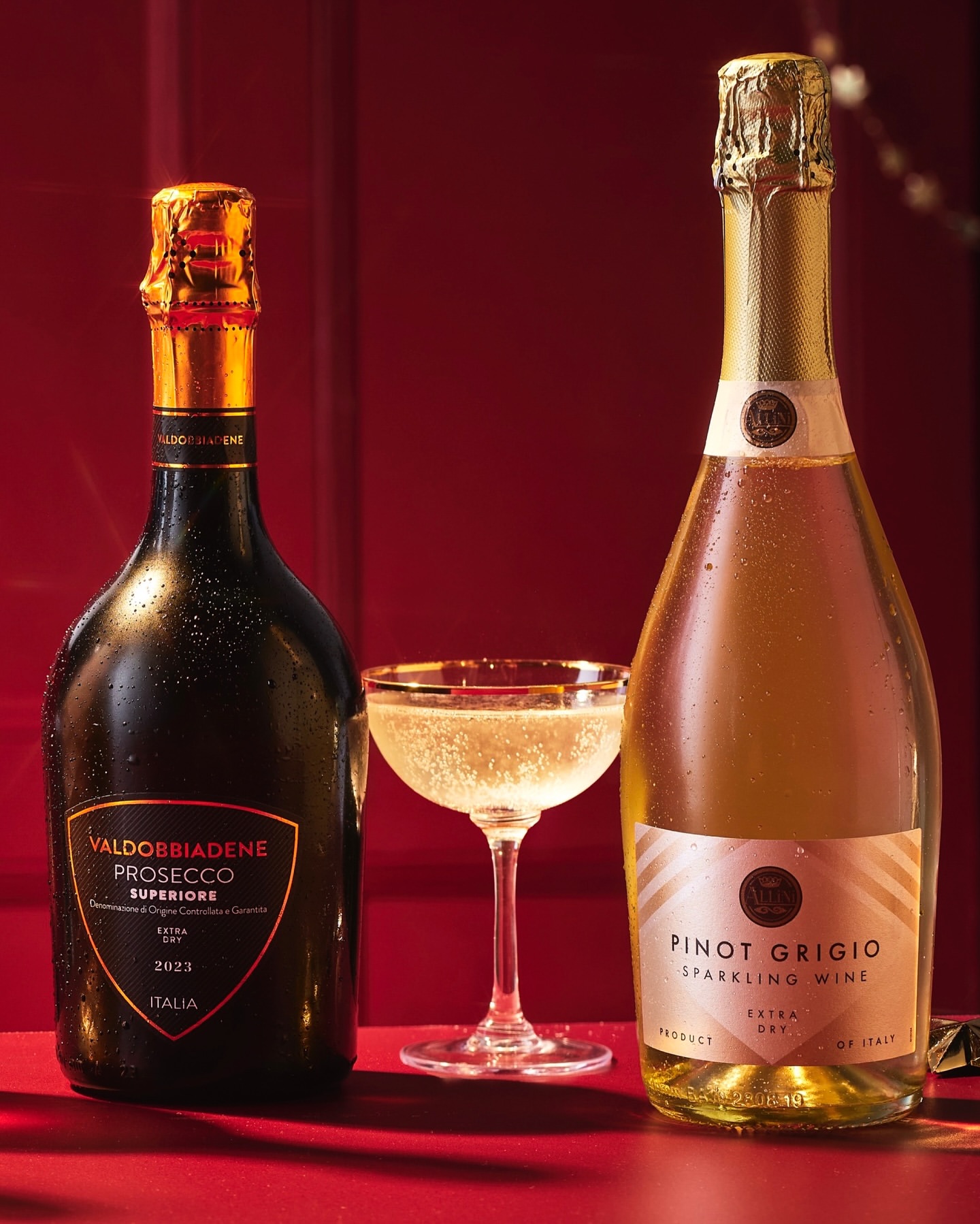 Two bottles of Prosecco and Pinot Grigio sparkling wine, with a full champagne coupe glass on a red surface against a red backdrop.