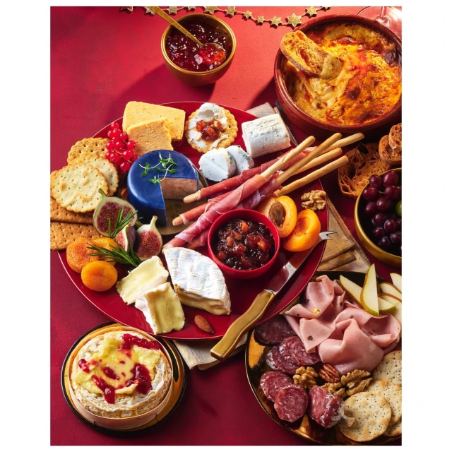Festive charcuterie board with various cheeses, meats, fruits, crackers, and accompaniments, presented on a red background.