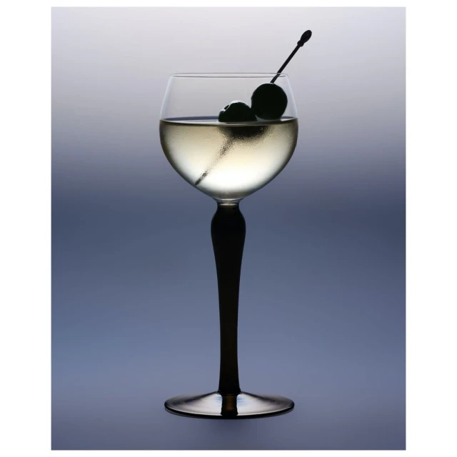 Elegant stemware glass with a clear liquid and garnished with two dark cherries against a blurred, graduated blue and grey background.