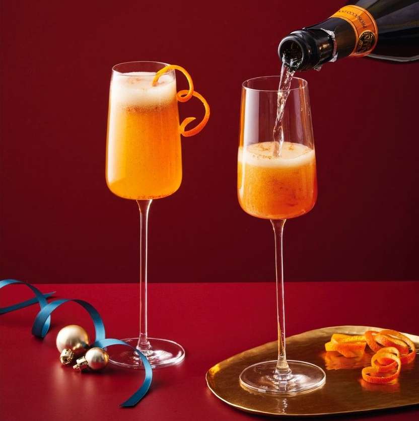 Two champagne flutes filled with mimosa, one being poured. Orange peel garnish and ribbon accent the drinks against a red backdrop.