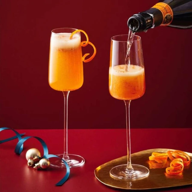 Two champagne flutes filled with mimosa, one being poured. Orange peel garnish and ribbon accent the drinks against a red backdrop.