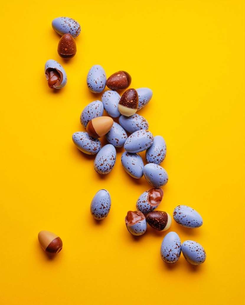 Overhead shot of blue, brown, and caramel chocolate candy eggs scattered on a bright yellow background, some broken open to reveal filling.