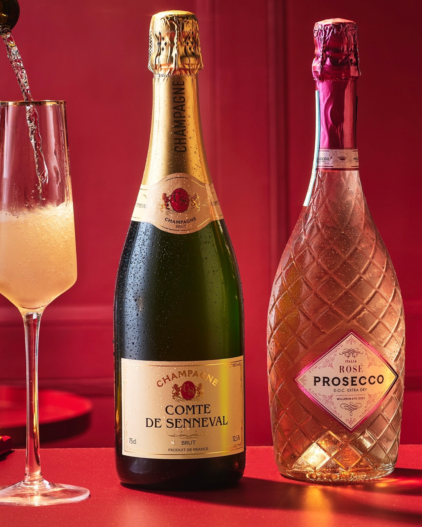 Two champagne bottles stand with a flute glass being filled with sparkling liquid, set against a vibrant red background for celebration.
