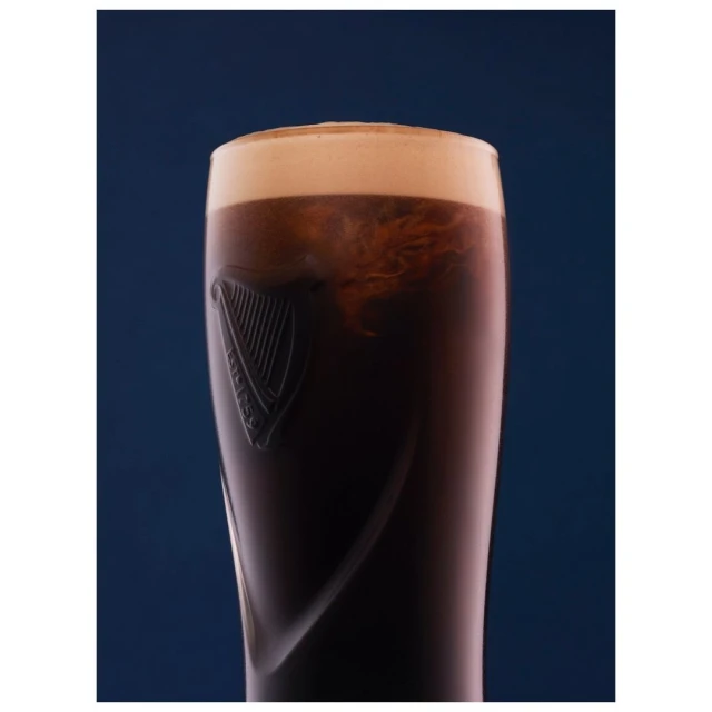 Close-up of a glass of dark stout beer with a creamy tan head, illuminated against a dark blue backdrop.