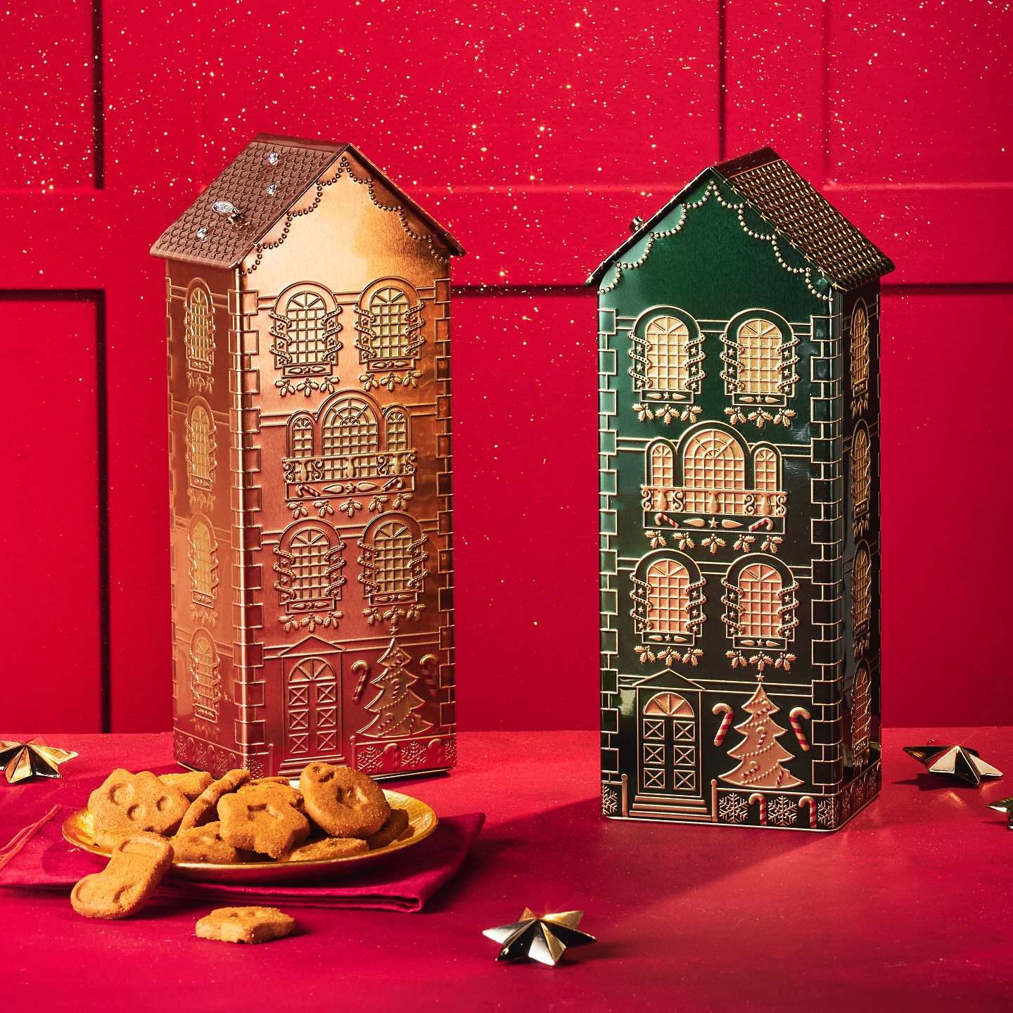 Two house-shaped metal tins with gingerbread cookies on a red surface, evoking a festive holiday treat and decorative packaging.