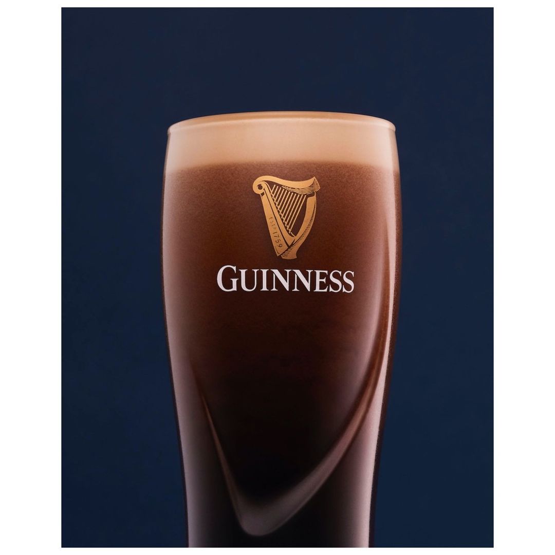 Close-up shot of a pint of Guinness beer featuring a creamy head, dark body, and brand logo, set against a blurred dark blue backdrop.