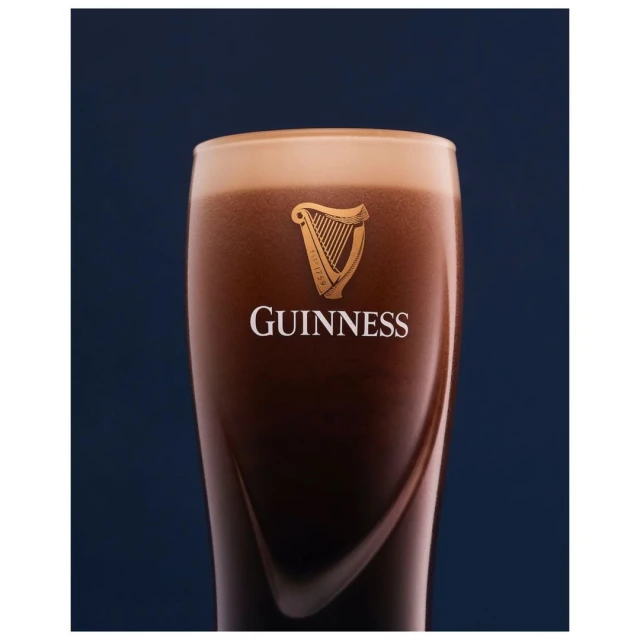 Close-up shot of a pint of Guinness beer featuring a creamy head, dark body, and brand logo, set against a blurred dark blue backdrop.