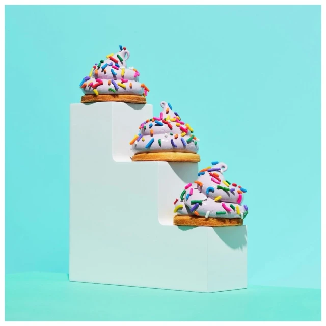 Three cookies with sprinkles are stacked on a white, three-step platform against an aqua background for a simple, colorful, studio shot.