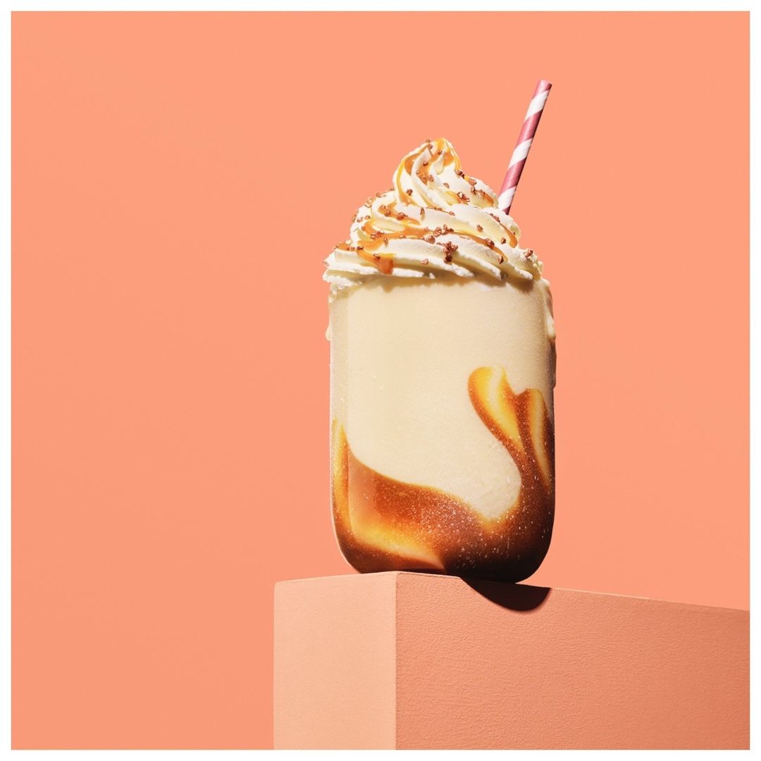 A creamy milkshake with whipped cream and caramel drizzle, topped with a pink striped straw, sits on a peach-colored cube against a peach background.