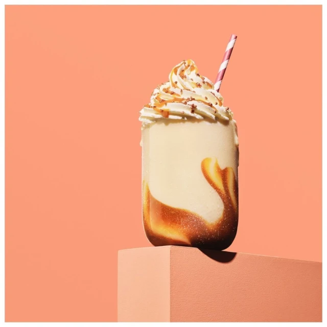 A creamy milkshake with whipped cream and caramel drizzle, topped with a pink striped straw, sits on a peach-colored cube against a peach background.