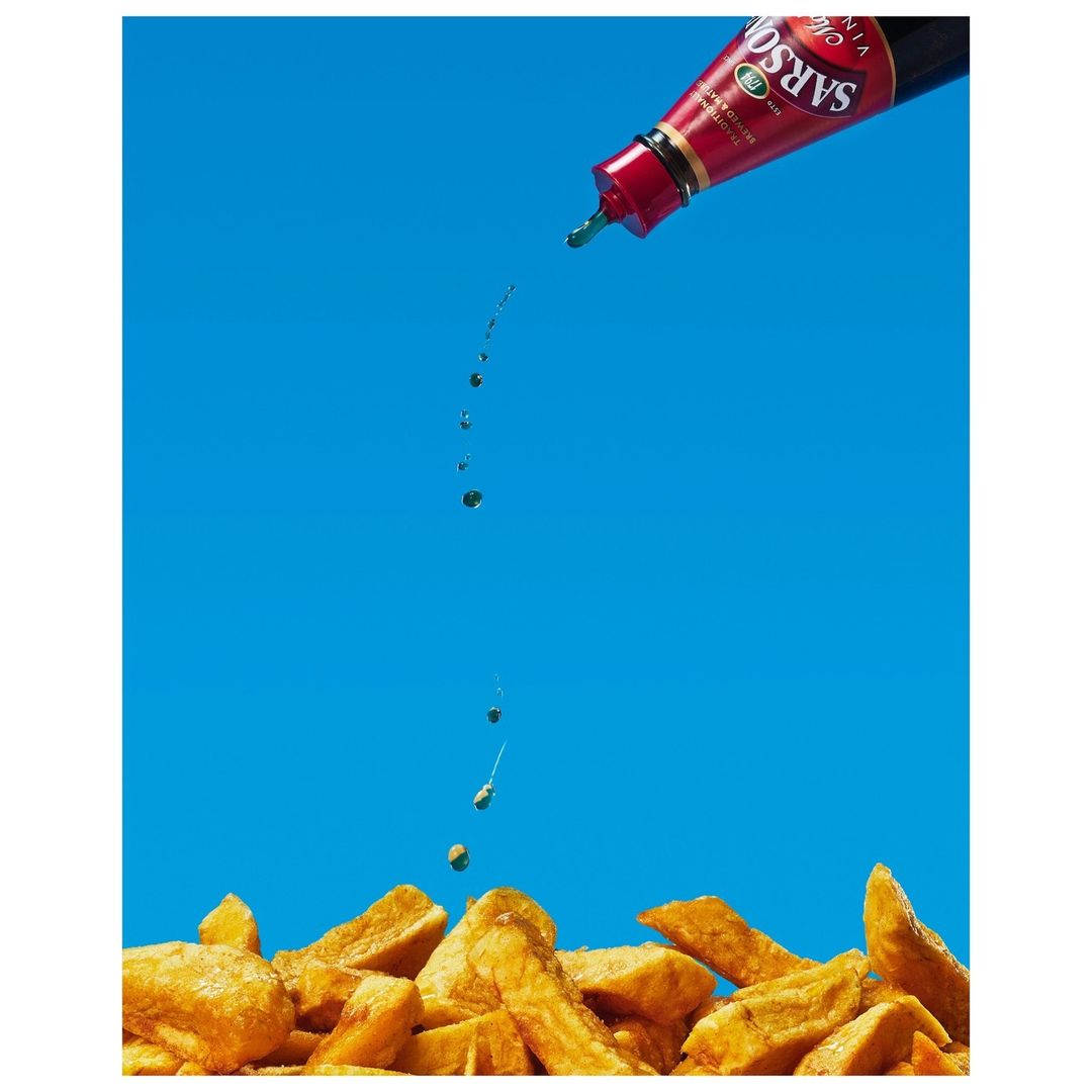 Pouring vinegar on french fries against a bright blue background. Still life image features a sauce bottle and golden fried chips.