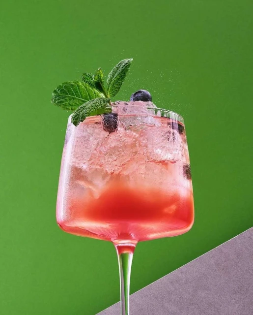 Refreshing mixed cocktail drink in a tall glass with ice, blueberries and a sprig of mint against green and grey backdrop.