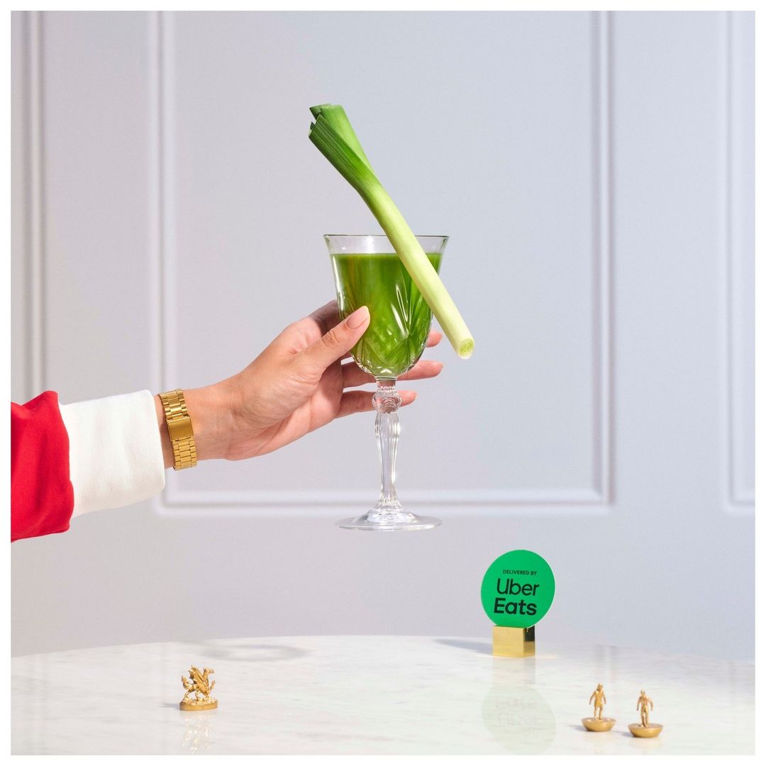 Hand holds a crystal glass with a green drink and a leek, with a tiny gold statue on the table and an Uber Eats logo card.