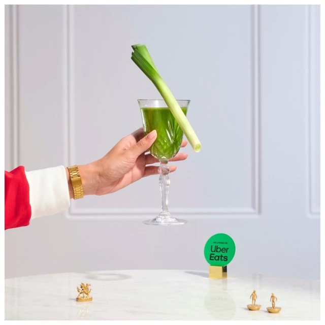 Hand holds a crystal glass with a green drink and a leek, with a tiny gold statue on the table and an Uber Eats logo card.