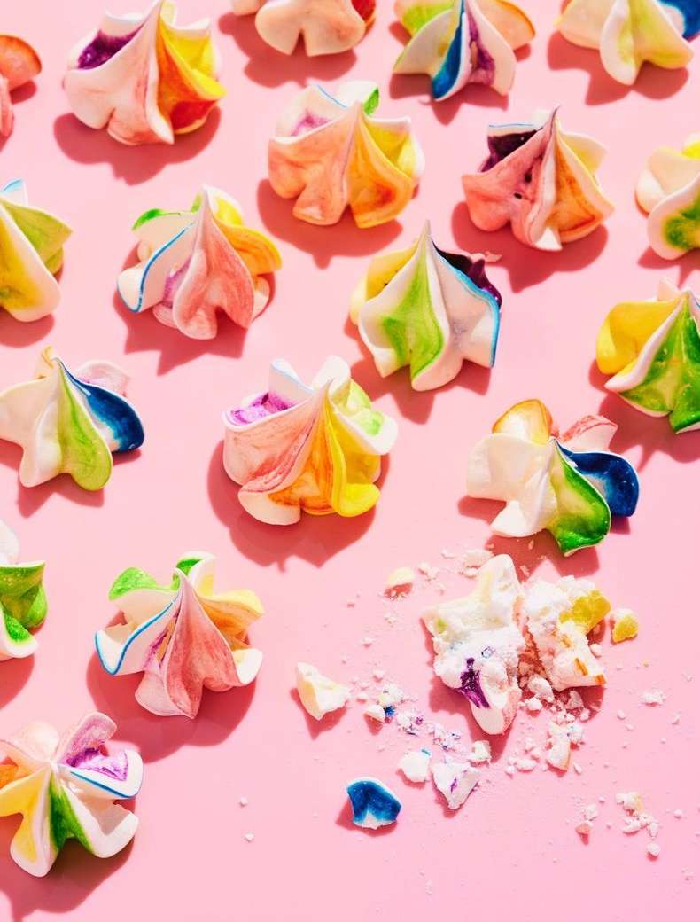Colorful meringue cookies arranged on a pink surface, some crushed, with bright, direct lighting creating soft shadows.