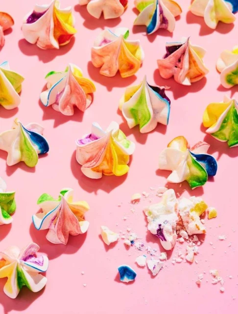 Colorful meringue cookies arranged on a pink surface, some crushed, with bright, direct lighting creating soft shadows.