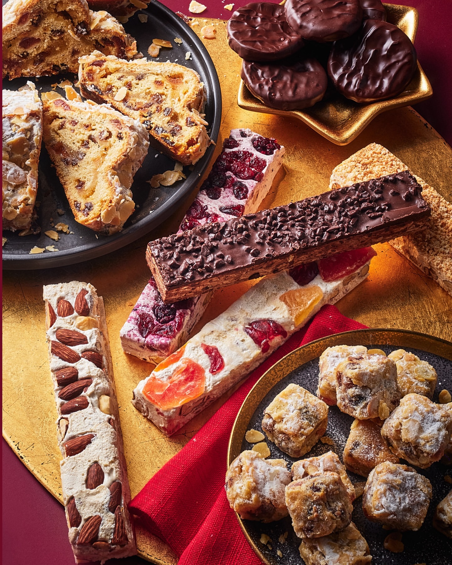 Assortment of festive treats, including stollen, nougat, and chocolate mints, displayed on plates with a gold and red backdrop.