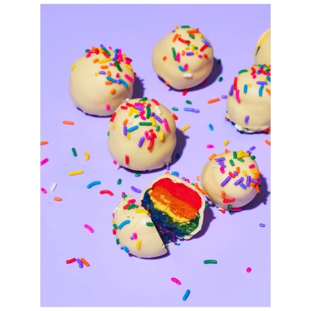 Seven rainbow-layer cake balls covered with white chocolate and sprinkles, displayed against a solid light purple background.