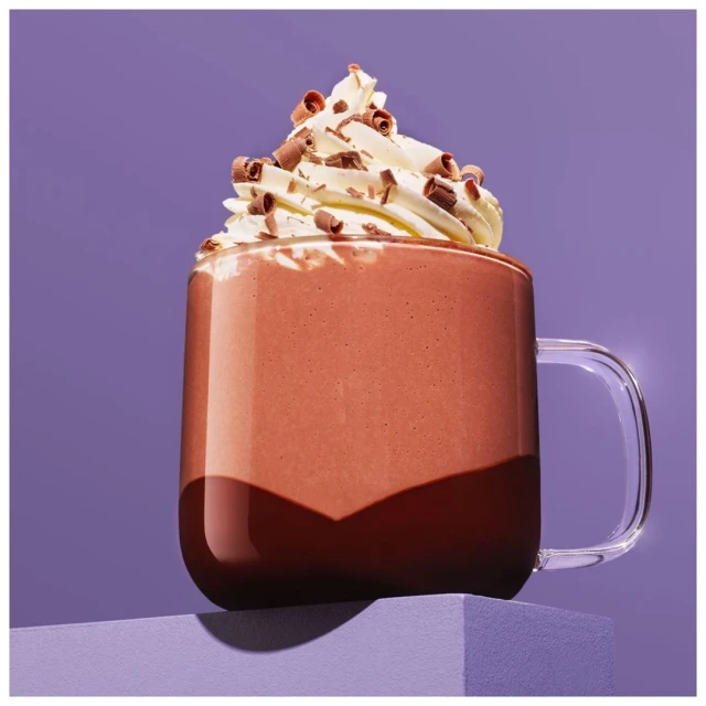 Hot chocolate in a glass mug with whipped cream and chocolate shavings, set on a purple stand against a light purple backdrop.