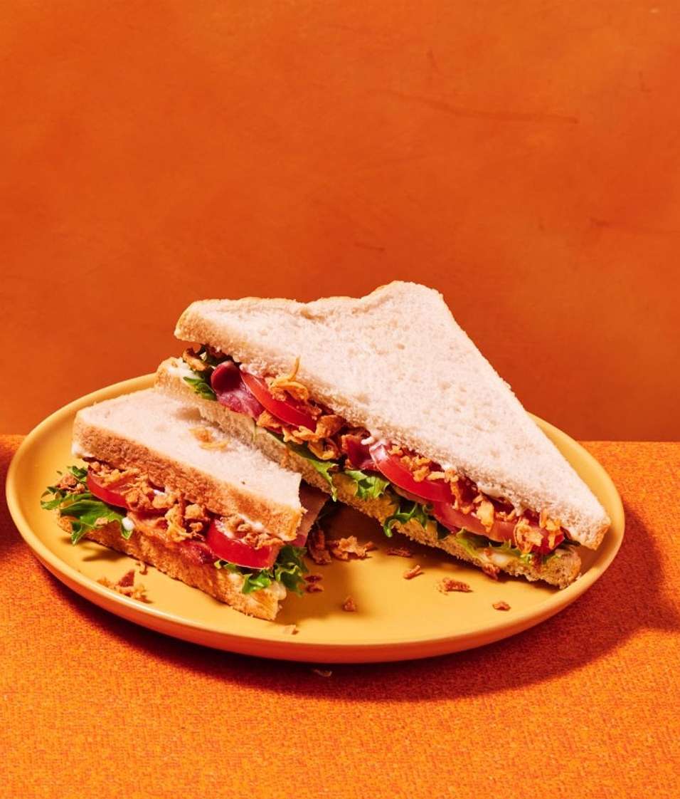 Overhead shot of two triangular sandwiches on a yellow plate, filled with lettuce, tomato, ham, and crispy fried onions against orange background.