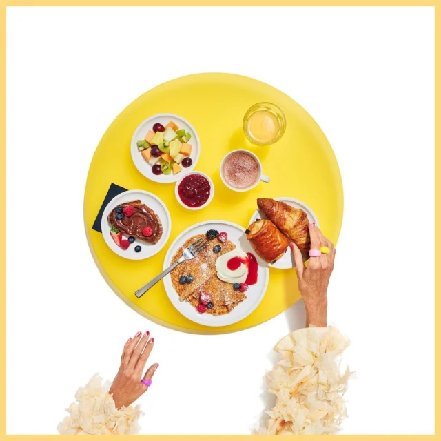 Overhead view of a yellow table laden with breakfast foods like pancakes, croissants, fruit, and jam, being served by two adult hands.