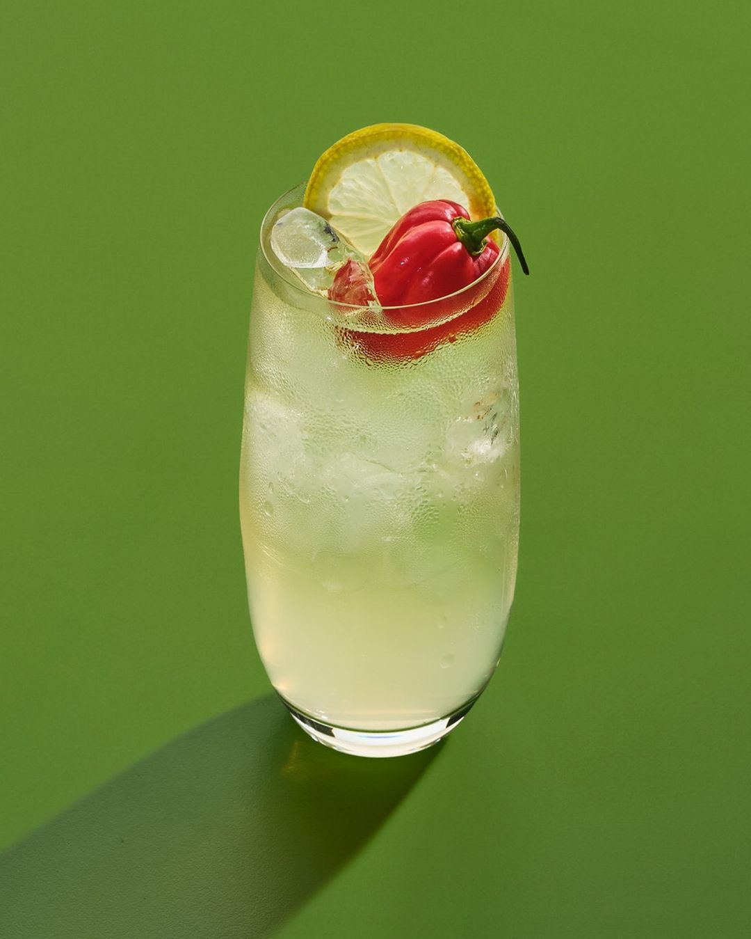 Refreshing cocktail in a tall glass with ice cubes, lemon slice, and habanero pepper garnish, presented on a solid green background.