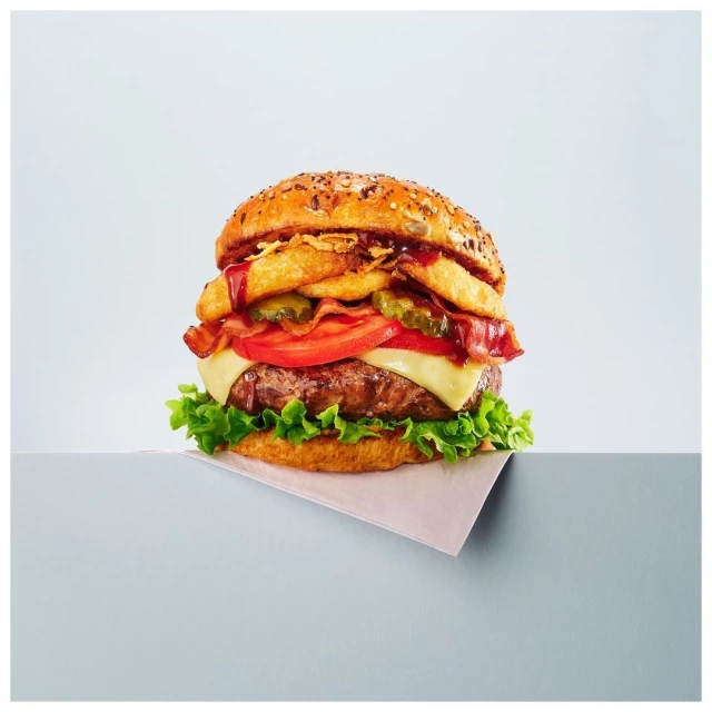 Close-up of a large hamburger with beef patty, cheese, vegetables, bacon, and onion rings, served on a grey surface against a grey background.