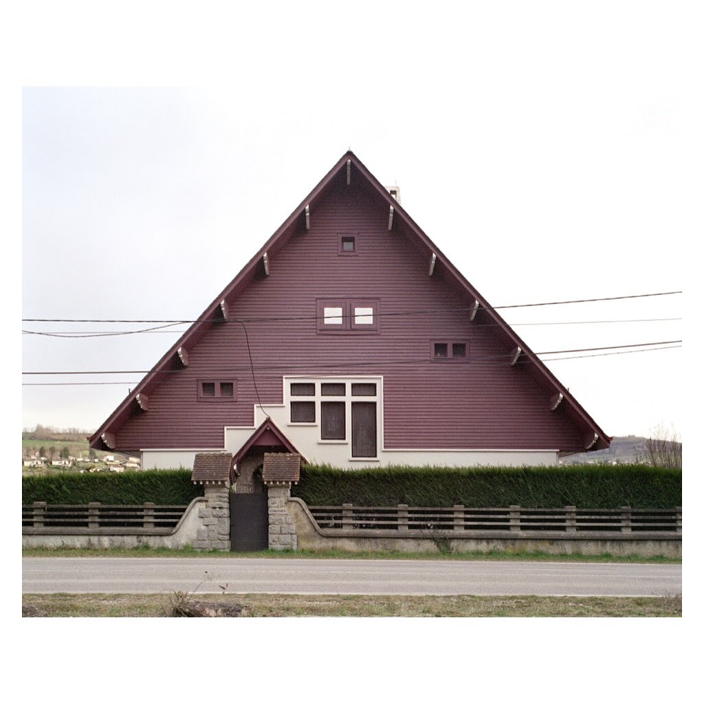 Exterior of a unique triangular house with burgundy siding, symmetrically framed with a gate, fence, and green hedges under an overcast sky.