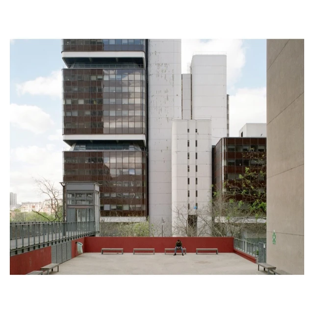 Person sitting on a bench on a red concrete terrace with modern high-rise buildings as backdrop under a partly cloudy sky during daytime.