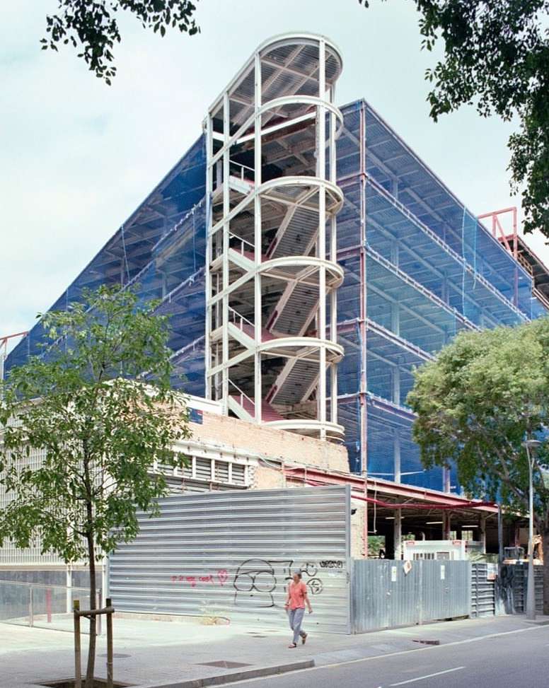 Outdoor shot of a modern building under construction with a scaffolding staircase, a passerby on the sidewalk, and a tree in the foreground.