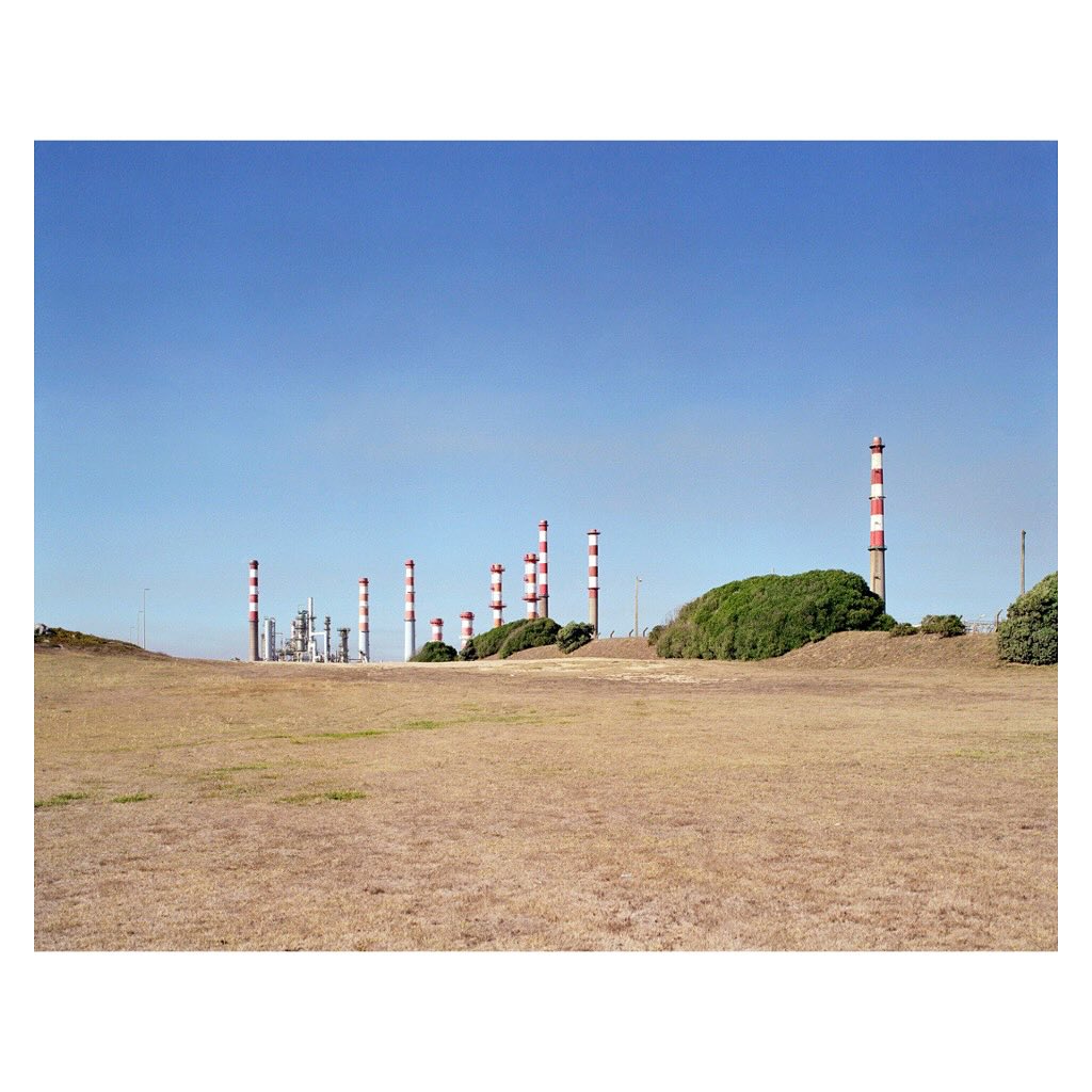 Industrial landscape featuring a vast field leading to a facility with multiple chimneys under a clear blue sky, creating a clean, minimalist feel.